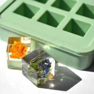 En grøn isterningbakke står ved siden af to klare isterninger, som hver indeholder farverige blomster - den ene med orange kronblade og den anden med små blå og grønne blomster - der er frosset ned indeni.